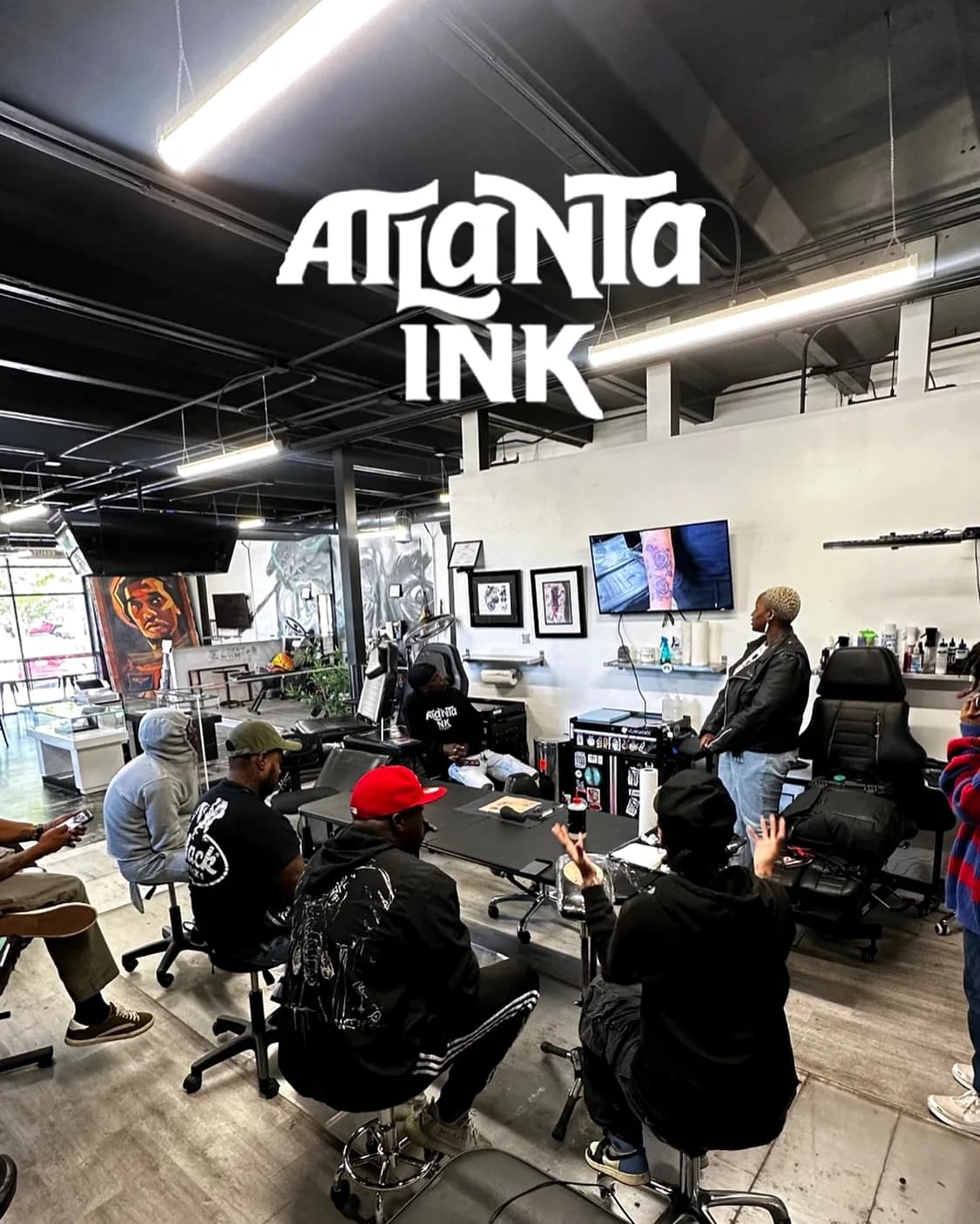 Staff and clients gathered around a table inside the ATLANTA INK studio with a tattoo chair and TV in the background
